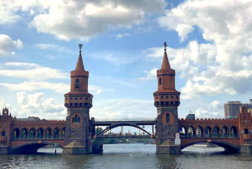 Die Bildbeschreibung zeigt die Oberbaumbrücke in Berlin, eine historische Brücke mit zwei markanten Türmen, die über einen Fluss führt. Im Hintergrund sind Wolken und einige Gebäude sichtbar, was die städtische Umgebung betont.