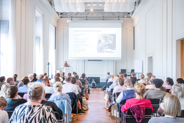 Eine Präsentation in einem hellen Raum mit vielen Sitzplätzen, wo eine Referentin vor einem Bildschirm steht. Das Publikum hört aufmerksam zu, während Informationen auf der Leinwand angezeigt werden. Die Atmosphäre ist fokussiert und professionell.