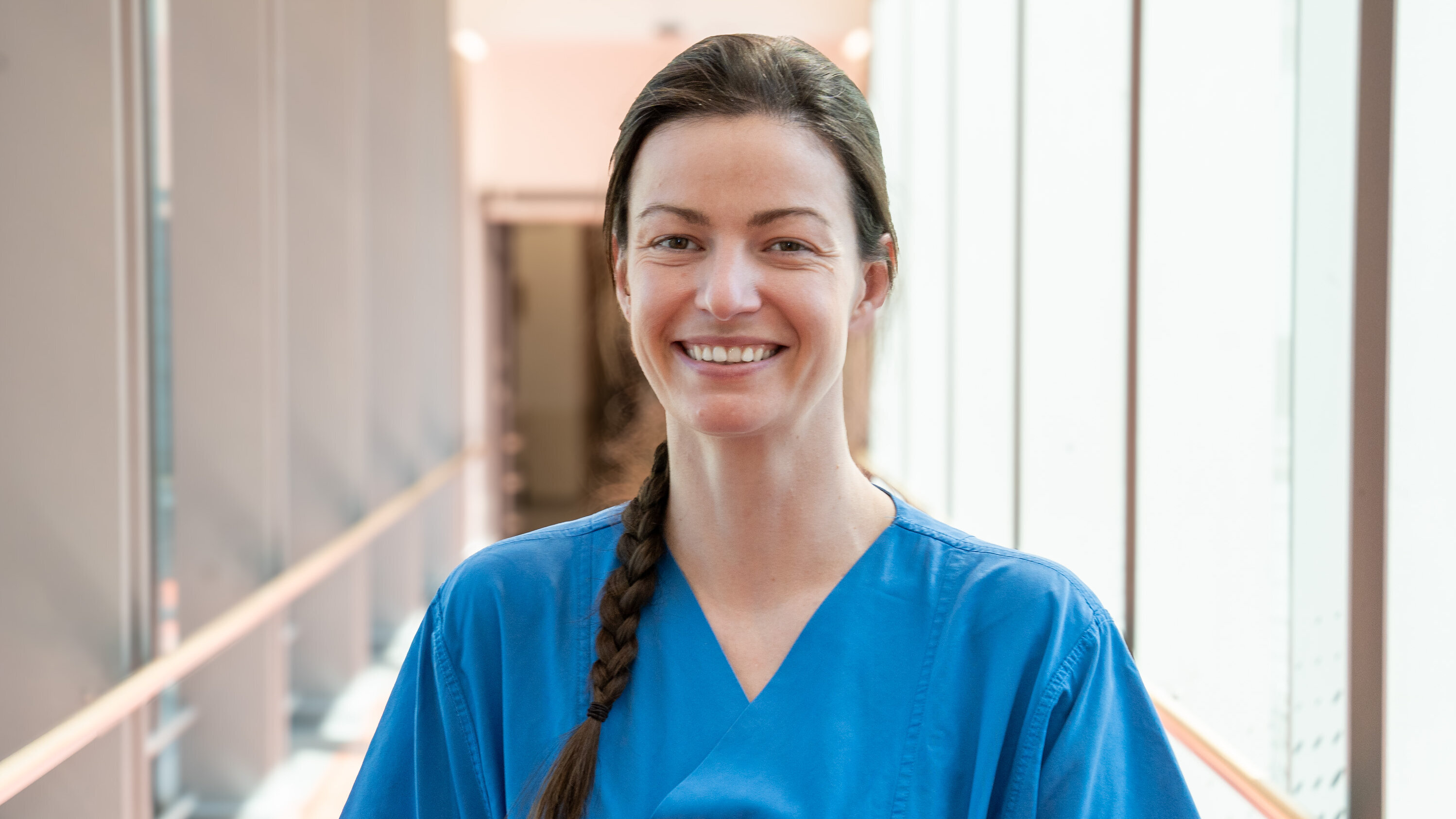 Eine lächelnde Frau in einem blauen medizinischen Outfit steht in einem hellen Flur. Ihr Haar ist zu einem langen Zopf geflochten. Sie vermittelt einen freundlichen und professionellen Eindruck.