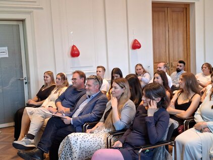 Eine Gruppe von Personen sitzt in einem Konferenzraum. Die Teilnehmer hören aufmerksam zu und sind unterschiedlich gekleidet. Im Hintergrund sind rote Dekorationen sichtbar. Die Atmosphäre wirkt konzentriert und informativ.