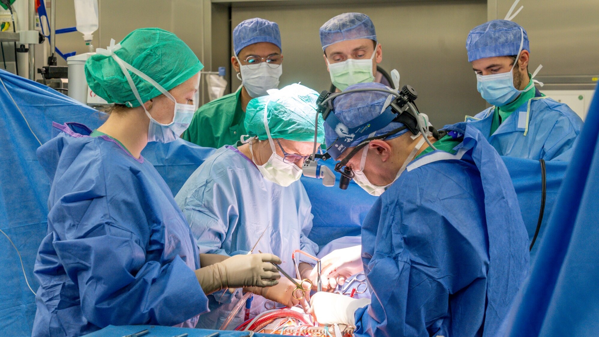 Sechs Personen in Operationskleidung stehen um einen Operationstisch. Zwei Chirurgen führen einen Eingriff durch, während ein Teamkollege sie unterstützt. Alle tragen Gesichtsmasken und Schutzbrillen. Instrumente liegen auf dem Tisch bereit, während konzentrierte Blicke auf dem Eingriff ruhen.