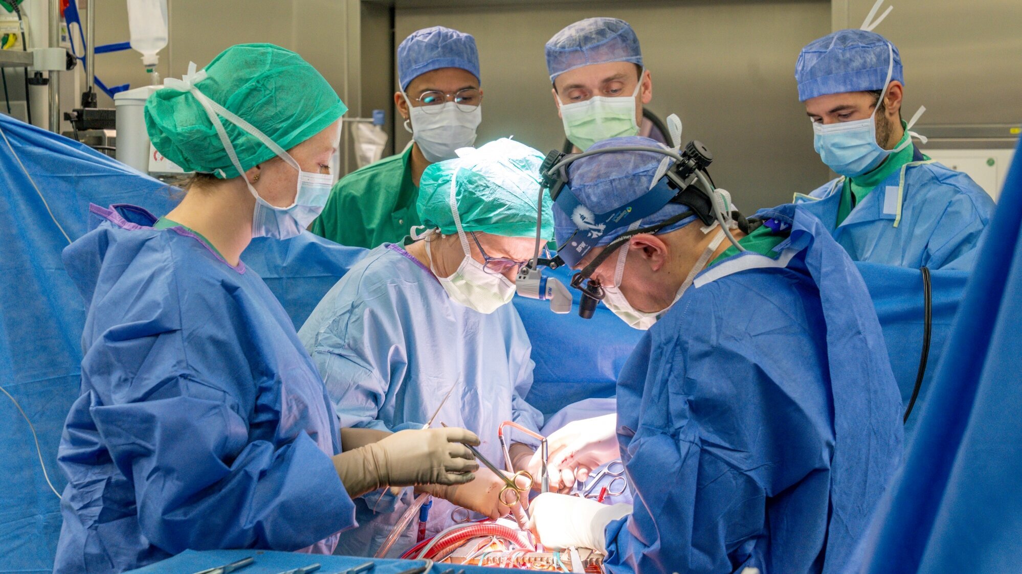 Sechs Personen in Operationskleidung stehen um einen Operationstisch. Zwei Chirurgen führen einen Eingriff durch, während ein Teamkollege sie unterstützt. Alle tragen Gesichtsmasken und Schutzbrillen. Instrumente liegen auf dem Tisch bereit, während konzentrierte Blicke auf dem Eingriff ruhen.