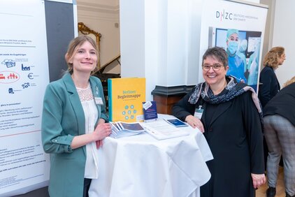 Zwei Frauen stehen an einem Stehtisch bei einer Veranstaltung. Eine von ihnen hält ein gelbes Buch mit dem Titel „Medizin Regelmappe“. Auf dem Tisch liegen Informationsmaterialien. Im Hintergrund sind Plakate und weitere Besucher der Veranstaltung zu sehen.