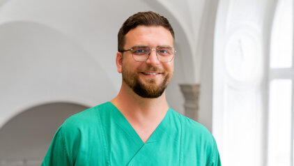 Ein lächelnder Mann trägt ein grünes medizinisches Outfit und Brille. Er steht in einem hellen Raum mit großen Fenstern im Hintergrund. Die Atmosphäre wirkt professionell und freundlich.