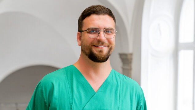 Ein lächelnder Mann trägt ein grünes medizinisches Outfit und Brille. Er steht in einem hellen Raum mit großen Fenstern im Hintergrund. Die Atmosphäre wirkt professionell und freundlich.