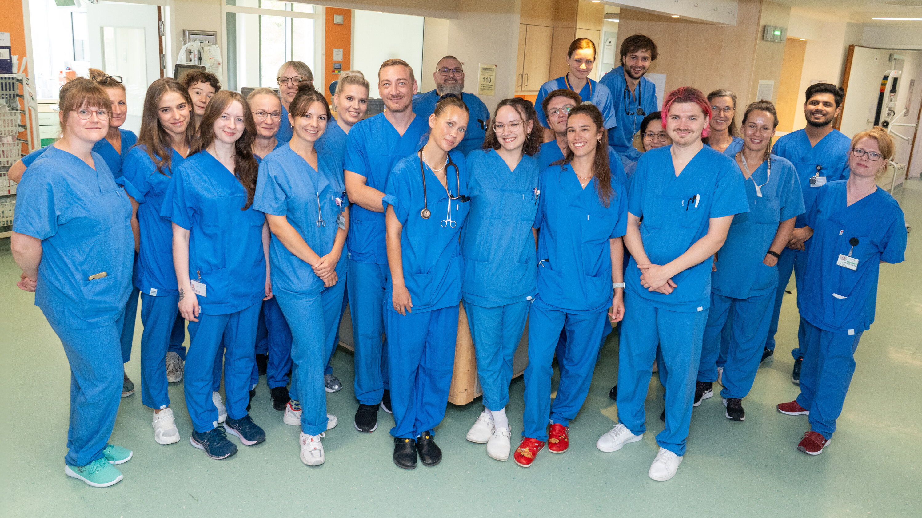 Eine Gruppe von 23 Personen in blauen Scrubs steht in einem Krankenhaus. Einige tragen Stethoskope. Sie lächeln und stehen eng zusammen, um den Fotografen anzusehen. Im Hintergrund sind medizinische Geräte und ein hell beleuchteter Raum sichtbar.