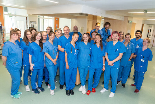 Eine Gruppe von 23 Personen in blauen Scrubs steht in einem Krankenhaus. Einige tragen Stethoskope. Sie lächeln und stehen eng zusammen, um den Fotografen anzusehen. Im Hintergrund sind medizinische Geräte und ein hell beleuchteter Raum sichtbar.