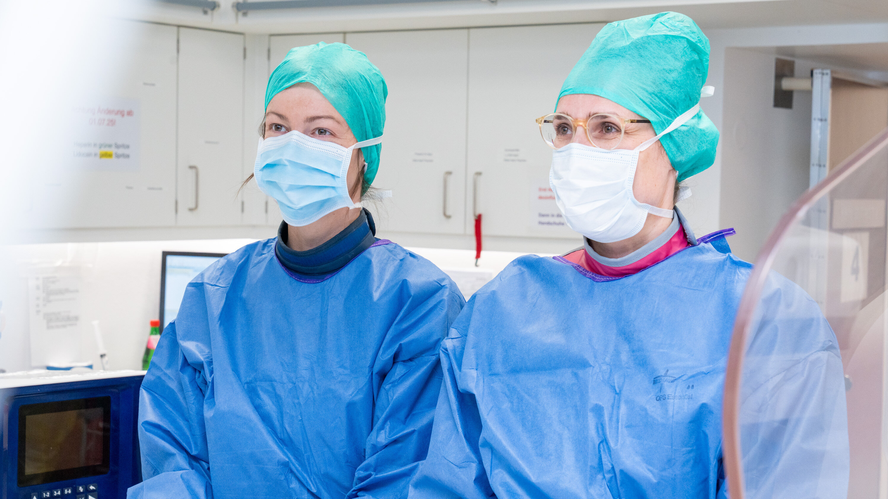 Zwei weibliche medizinische Fachkräfte tragen Schutzkleidung, chirurgische Masken und Hauben. Sie stehen in einem modernen, hellen Krankenhausraum und blicken aufmerksam. Im Hintergrund sind medizinische Geräte und eine sterile Umgebung zu erkennen.