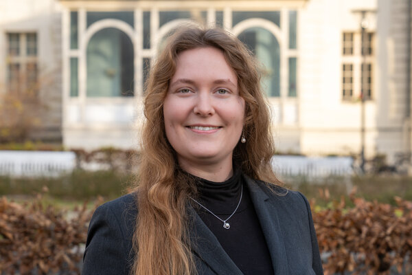 Ein lächelnde Frau mit langen, lockigen Haaren trägt ein schwarzes Oberteil und einen grauen Blazer. Sie steht vor einem historischen Gebäude mit großen Fenstern. Im Hintergrund sind trockene Pflanzen sichtbar.