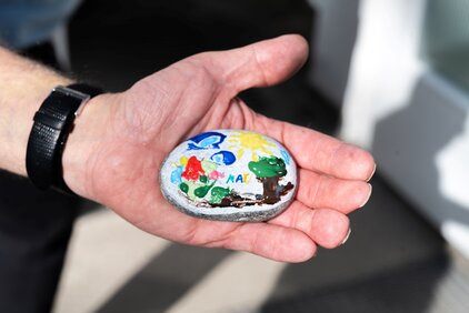 Eine Hand hält einen bemalten Stein. Der Stein zeigt eine bunte Darstellung mit grünen Bäumen, einer Sonne und roten Blumen. Auf dem Stein steht das Wort "MUT" in blauer Schrift.