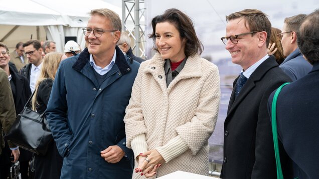 Drei Personen stehen in einem informellen Moment zusammen, lächelnd und freundlich. Sie tragen legere, moderne Kleidung und befinden sich anscheinend auf einer Veranstaltung im Freien. Im Hintergrund sind unscharfe Silhouetten von weiteren Menschen und einem Zelt zu sehen.