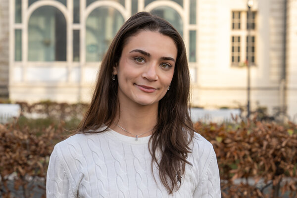 Eine junge Frau mit langen, braunen Haaren lächelt in die Kamera. Sie trägt einen hellen, gestrickten Pullover und eine dezente Halskette. Im Hintergrund sind verschwommene, lichtdurchflutete Gebäude und Pflanzen sichtbar.