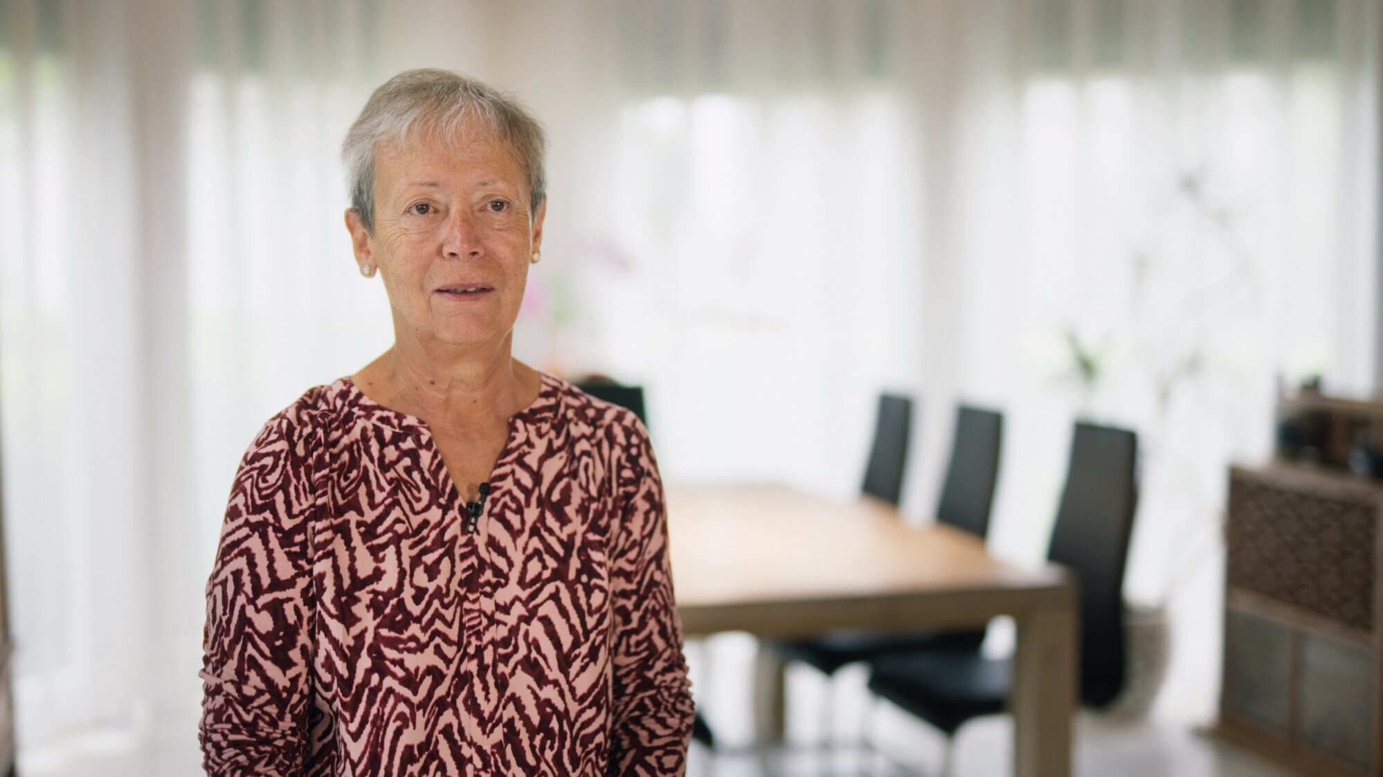 Eine ältere Frau mit kurzen, grauen Haaren steht in einem hellen Raum. Sie trägt ein gemusternes Oberteil und lächelt leicht. Im Hintergrund ist ein Esszimmer mit einem Tisch und Stühlen zu sehen. Das Licht fällt durch die Fenster und schafft eine freundliche Atmosphäre.