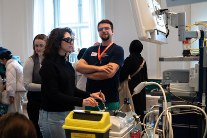 Eine Person mit einer speziellen Brille arbeitet an einem medizinischen Gerät, während ein Mann im Hintergrund zusieht. Weitere Personen stehen in der Nähe, in einem Raum mit modernen medizinischen Technologien und einem großen Bildschirm.