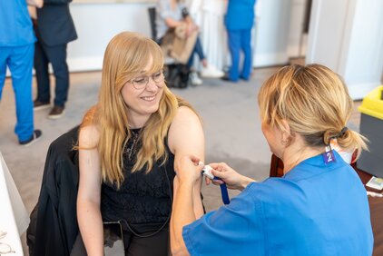 Eine Frau mit langen, blonden Haaren sitzt lächelnd, während eine medizinische Fachkraft ihr eine Spritze in den Arm setzt. Beide tragen entspannte Gesichtsausdrücke in einer hellen Umgebung. Im Hintergrund sind weitere Personen in blauen Kitteln zu sehen.