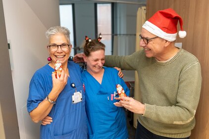 Drei Personen in blauen Arbeitsuniformen lächeln und halten Schokoladenfiguren in Form von Weihnachtsmännern. Eine Person trägt einen roten Weihnachtsmannhut. Die Gruppe scheint fröhlich und feiert gemeinsam in einem freundlichen, festlichen Ambiente.