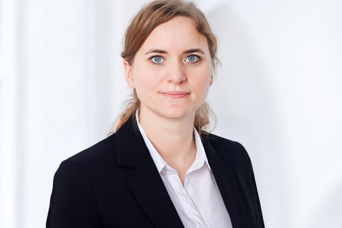 Eine junge Frau mit blonden Haaren und blauen Augen trägt einen schwarzen Blazer und ein weißes Hemd. Sie schaut direkt in die Kamera und hat einen freundlichen, selbstbewussten Ausdruck. Der Hintergrund ist hell und unscharf, um den Fokus auf ihr Gesicht zu legen.