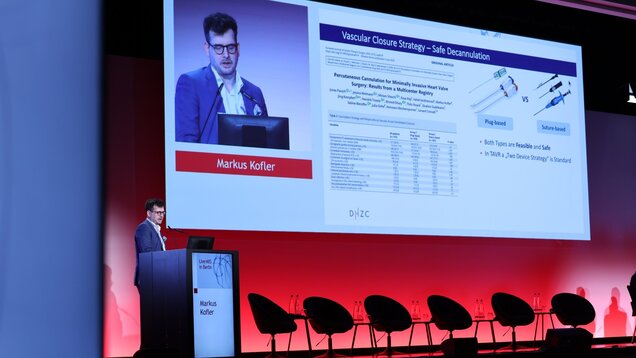 Ein Mann steht an einem Podium und präsentiert eine Folie über "Vaskuläre Verschlussstrategie - Sichere Dekanulation". Die Folie zeigt eine Tabelle mit Vergleichsdaten und zwei Bilder von medizinischen Instrumenten. Im Hintergrund sind Stühle und eine rote Lichtinstallation zu sehen.