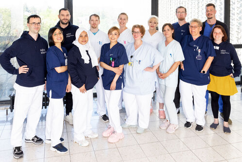 Eine Gruppe von 15 Fachkräften in medizinischer Kleidung steht lächelnd zusammen. Sie tragen unterschiedliche Oberteile in Blau und Weiß. Der Hintergrund zeigt Fenster mit Tageslicht, das eine angenehme Atmosphäre schafft. Die Menschen wirken freundlich und engagiert.