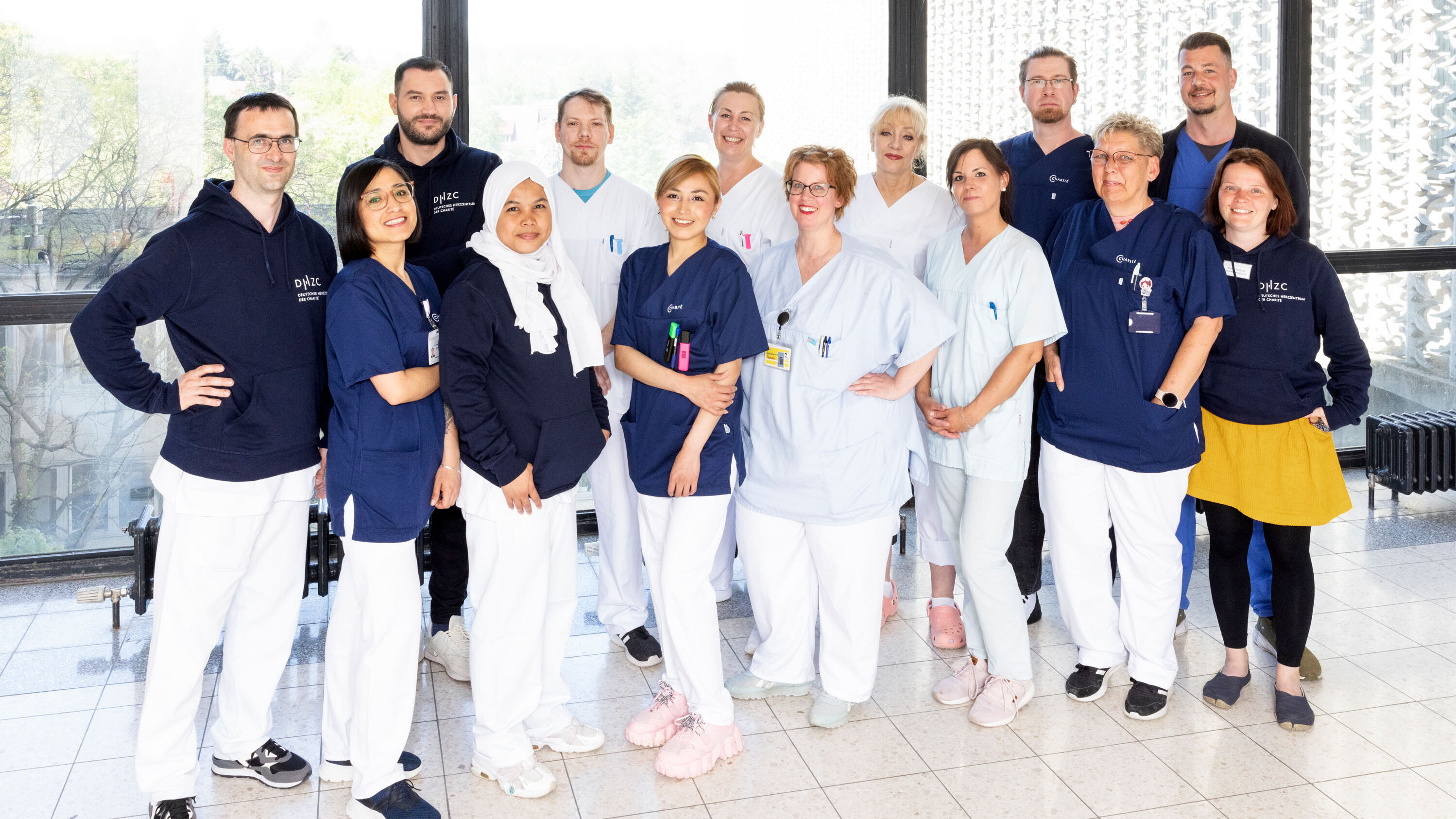 Eine Gruppe von 15 Fachkräften in medizinischer Kleidung steht lächelnd zusammen. Sie tragen unterschiedliche Oberteile in Blau und Weiß. Der Hintergrund zeigt Fenster mit Tageslicht, das eine angenehme Atmosphäre schafft. Die Menschen wirken freundlich und engagiert.