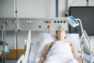 Ein Simulationsmodell liegt auf einem Krankenhausbett, bedeckt mit einem Bettlaken. Im Hintergrund sind medizinische Geräte und Monitore sichtbar, die für die Patientenüberwachung verwendet werden. Die Szene vermittelt eine klinische Umgebung zur Ausbildung oder Demonstration im Gesundheitswesen.