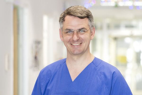 Ein männlicher Arzt in blauer Arbeitskleidung lächelt freundlich in die Kamera. Im Hintergrund sind unscharfe Krankenhausflure zu sehen. Der Arzt trägt eine Brille und hat kurzes, graues Haar.