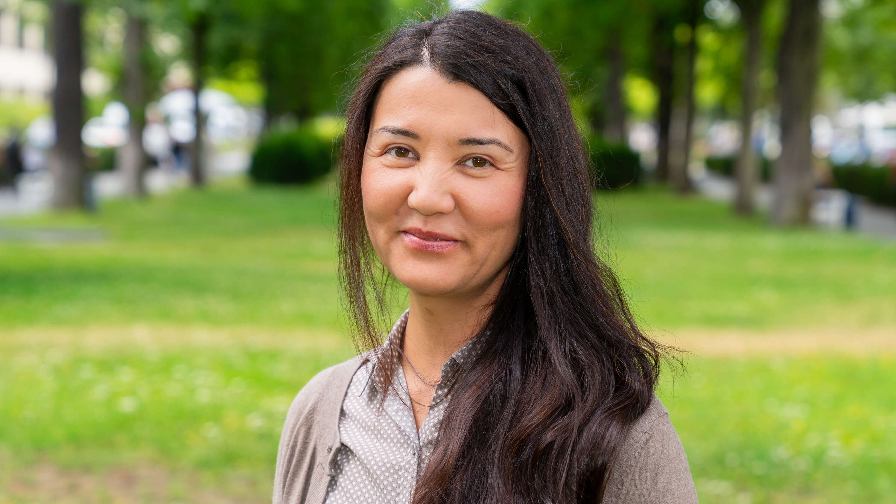 Eine Frau mit langen, dunklen Haaren lächelt in die Kamera. Sie trägt ein graues Oberteil mit einer hellen Bluse. Im Hintergrund ist eine grüne Wiese mit Bäumen und verschwommenen Menschen zu sehen. Die Szene vermittelt eine freundliche und entspannte Atmosphäre.
