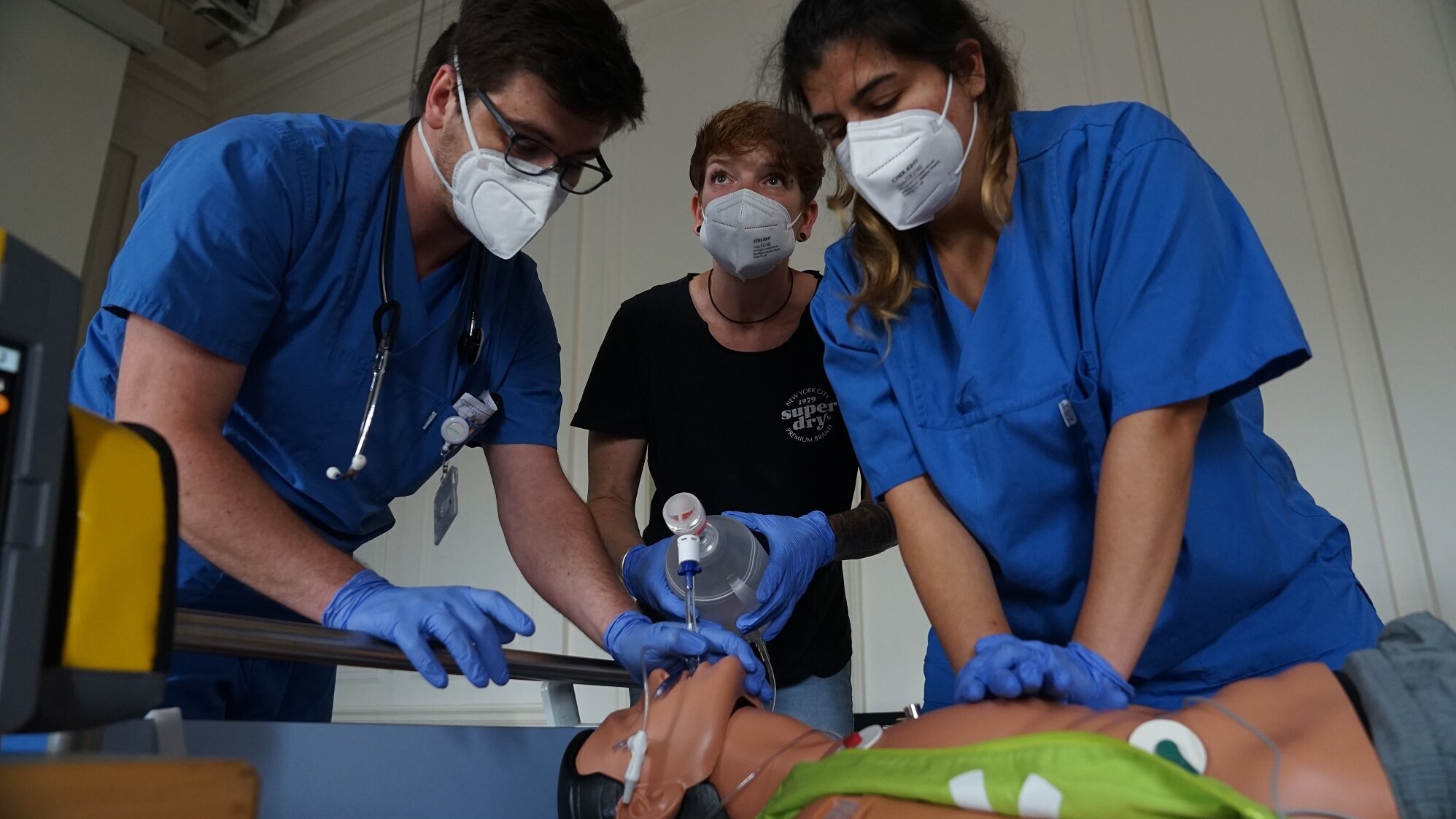 Drei Personen in blauen medizinischen Kitteln üben an einer Reanimationspuppe. Zwei Personen führen Atem- und Herzmassage durch, während eine dritte die Technik beobachtet. Alle tragen Schutzmasken.