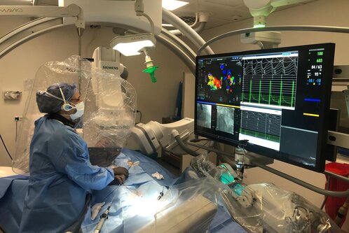 Ein Arzt in Schutzkleidung steht in einem modernen Operationssaal. Er arbeitet an einem Patienten, der auf einem Tisch liegt, umgeben von medizinischen Geräten. Ein großer Monitor zeigt farbige medizinische Grafiken und Vitalfunktionen an, während der Arzt ein präzises Verfahren durchführt.