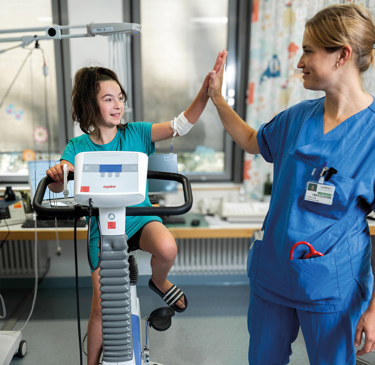 Ein Kind sitzt auf einem Ergometer und gibt einer Krankenschwester einen High-Five. Die Krankenschwester trägt einen blauen Kittel und lächelt. Im Hintergrund sind medizinische Geräte und eine helle, freundliche Umgebung zu sehen.