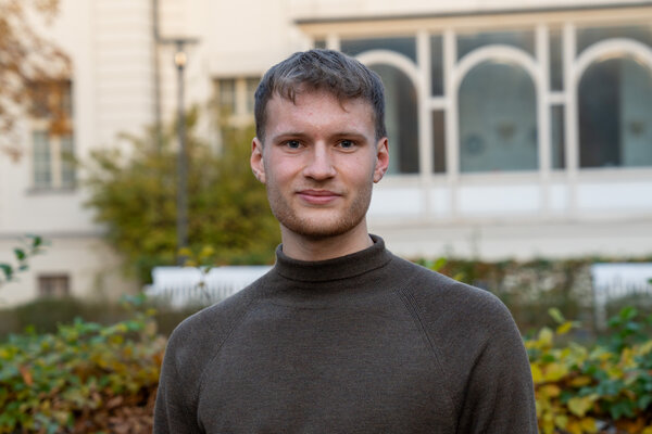 Ein junger Mann mit kurzen, grauen Haaren trägt einen dunklen Pullover und steht lächelnd vor einer bewachsenen Umgebung mit Sträuchern und Bäumen. Im Hintergrund sind Fenster und eine helle Gebäudefassade sichtbar.