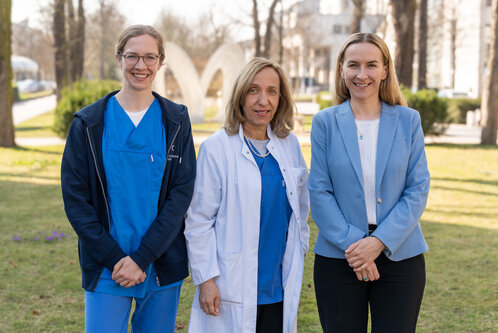 Drei Frauen stehen im Freien auf einer Wiese. Die erste trägt einen blauen Krankenhauskittel, die zweite einen weißen Arztkittel und die dritte ein hellblaues Sakko. Sie lächeln und präsentieren sich freundlich vor einer grünen Umgebung mit Bäumen im Hintergrund.