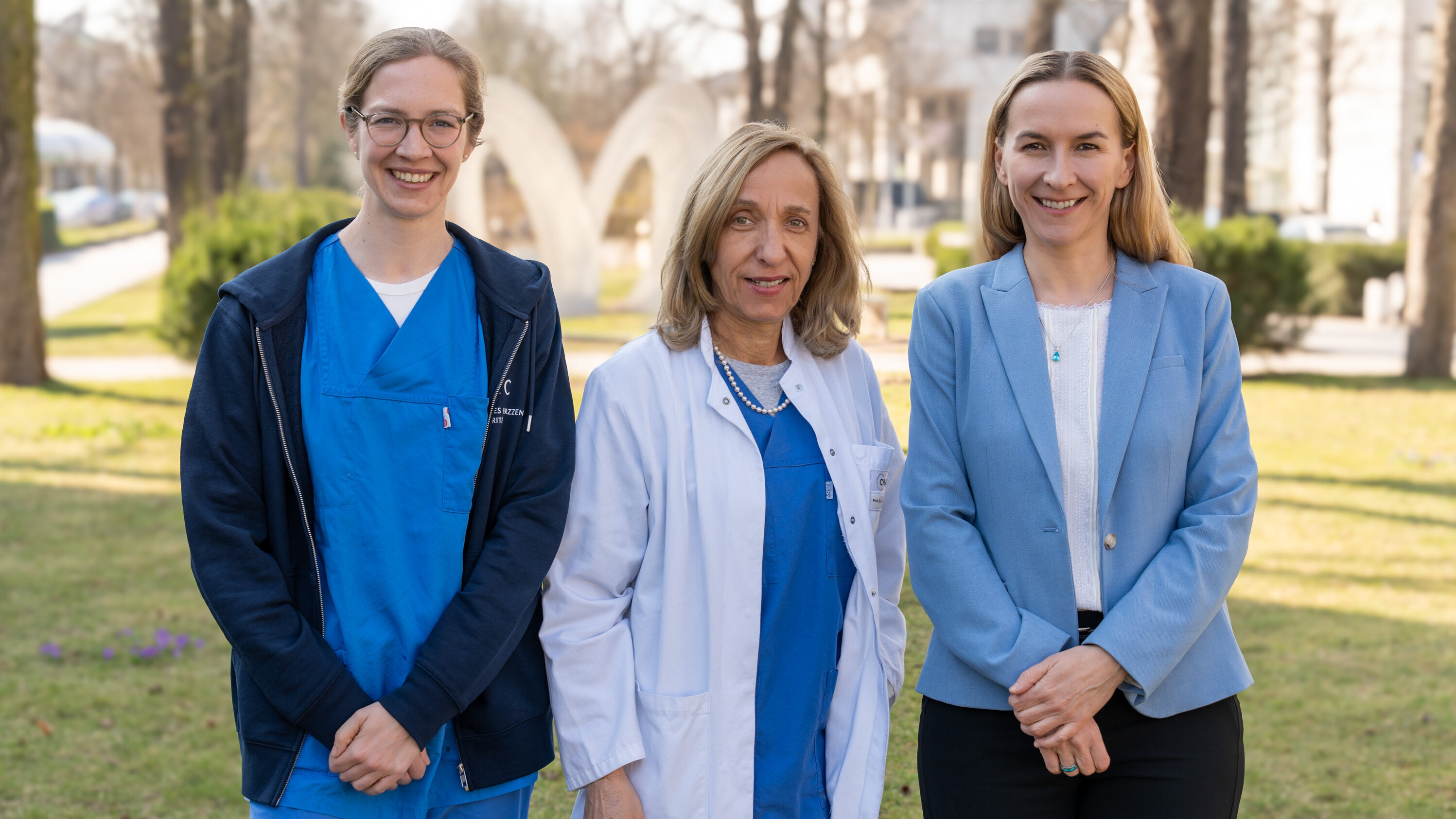 Drei Frauen stehen im Freien auf einer Wiese. Die erste trägt einen blauen Krankenhauskittel, die zweite einen weißen Arztkittel und die dritte ein hellblaues Sakko. Sie lächeln und präsentieren sich freundlich vor einer grünen Umgebung mit Bäumen im Hintergrund.