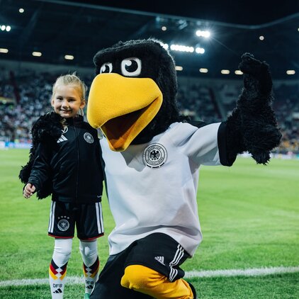 Ein Vereinsmaskottchen in Schwarz und Gelb steht auf dem Fußballplatz neben einem Mädchen, das ein Outfit in den Farben der deutschen Nationalmannschaft trägt. Beide lächeln und posieren fröhlich vor einer großen Menschenmenge im Hintergrund.
