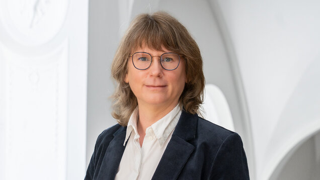 Porträt einer Person mit mittellangen, lockigen, braunen Haaren und Brille. Sie trägt einen dunkelblauen Blazer und ein weißes Hemd. Der Hintergrund zeigt einen hellen, architektonischen Raum mit runden Fenstern.