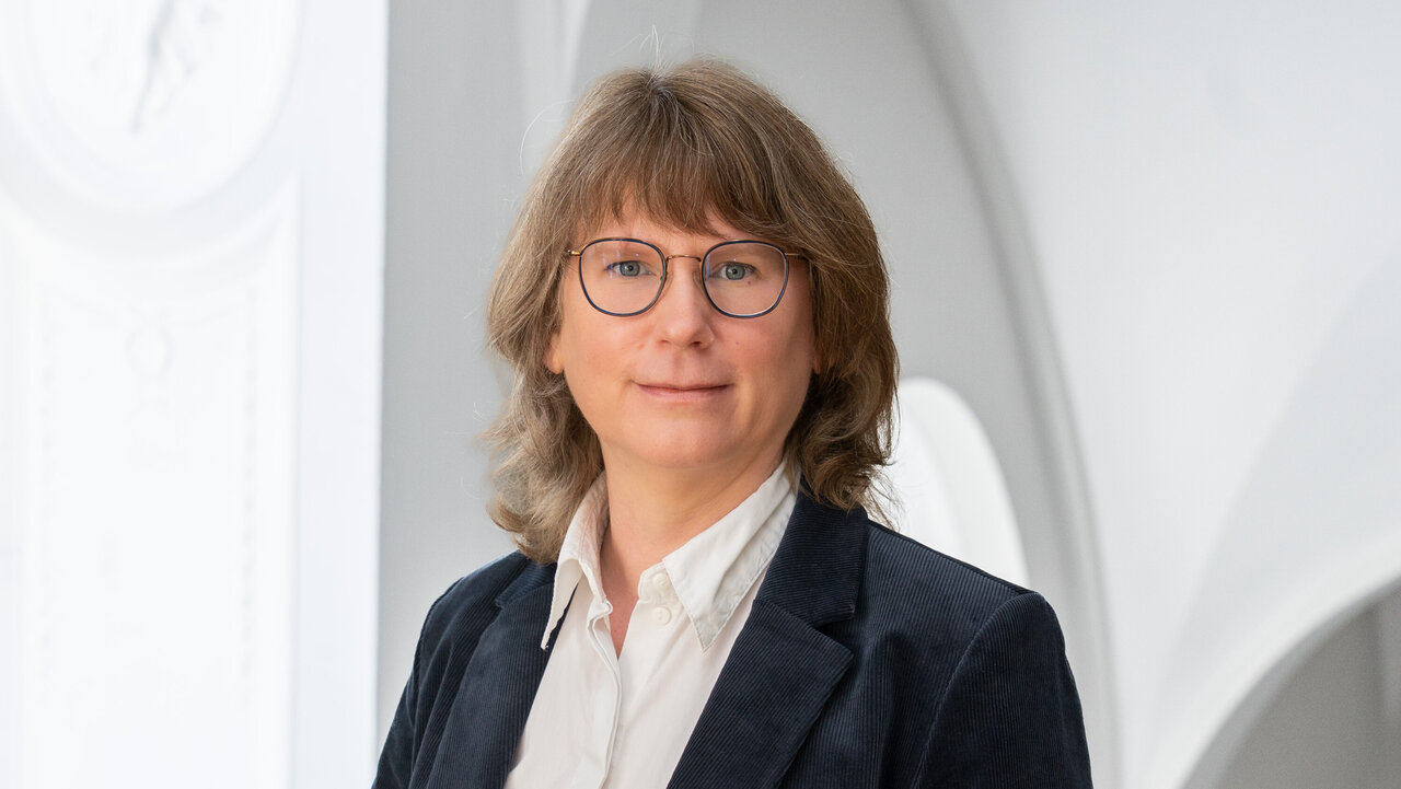 Porträt einer Person mit mittellangen, lockigen, braunen Haaren und Brille. Sie trägt einen dunkelblauen Blazer und ein weißes Hemd. Der Hintergrund zeigt einen hellen, architektonischen Raum mit runden Fenstern.