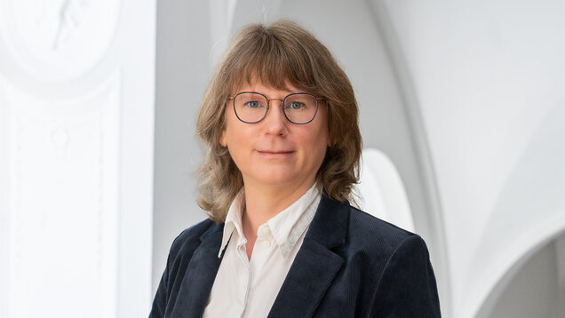 Porträt einer Person mit mittellangen, lockigen, braunen Haaren und Brille. Sie trägt einen dunkelblauen Blazer und ein weißes Hemd. Der Hintergrund zeigt einen hellen, architektonischen Raum mit runden Fenstern.
