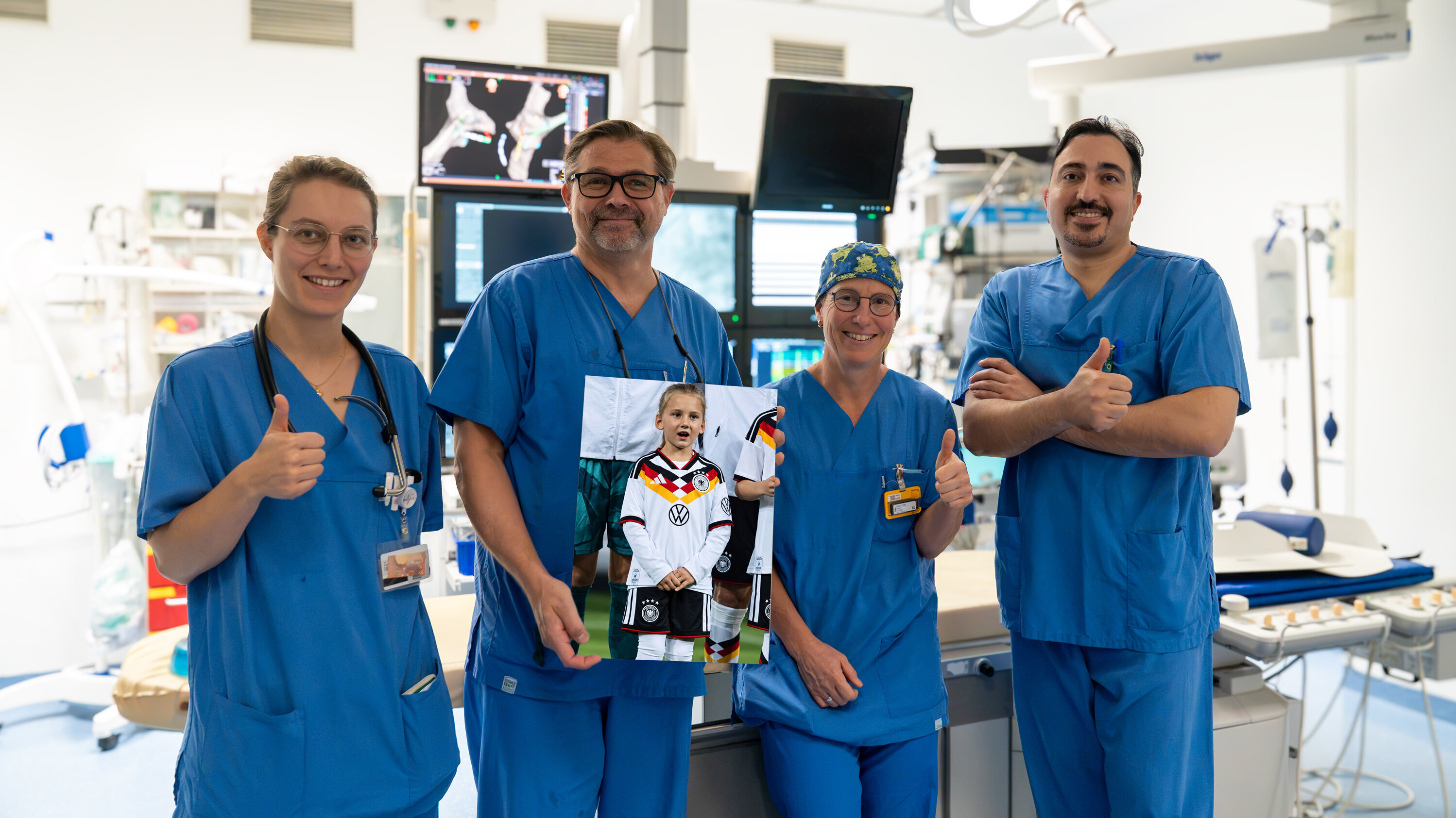 Vier medizinische Fachkräfte in blauer Kleidung stehen in einem Operationssaal. Sie lächeln und zeigen mit Daumen hoch auf ein Bild eines kleinen Mädchens, das in einem Fußballtrikot gekleidet ist. Der Hintergrund zeigt medizinische Geräte und Monitorbildschirme.