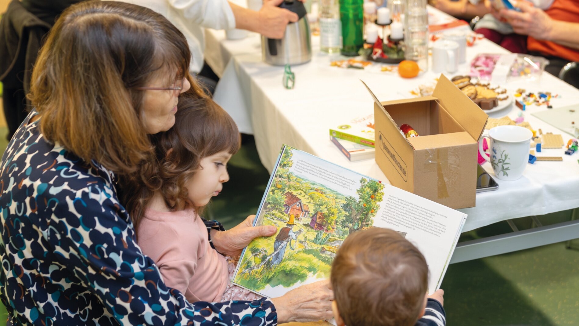 Eine erwachsene Frau sitzt mit zwei kleinen Kindern und zeigt ihnen ein illustriertes Buch. Die Kinder schauen interessiert auf die Seiten. Um sie herum sind Tische mit verschiedenen Gegenständen und Snacks.