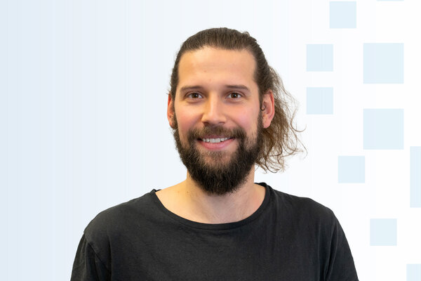 Ein junger Mann mit einem langen, lockigen Bart und langen, ungekämmten Haaren lächelt in die Kamera. Er trägt ein schwarzes T-Shirt und hat einen freundlichen Ausdruck. Der Hintergrund ist hellblau mit einfachen grafischen Elementen.