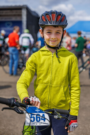 Ein Junge mit einem grünen Sweatshirt und einem Fahrradhelm steht lächelnd neben seinem Fahrrad. Sein Fahrrad hat eine Startnummer mit der Kennzeichnung "564". Im Hintergrund sind verschwommene Konturen von anderen Fahrradfahrern und Zuschauern zu sehen.