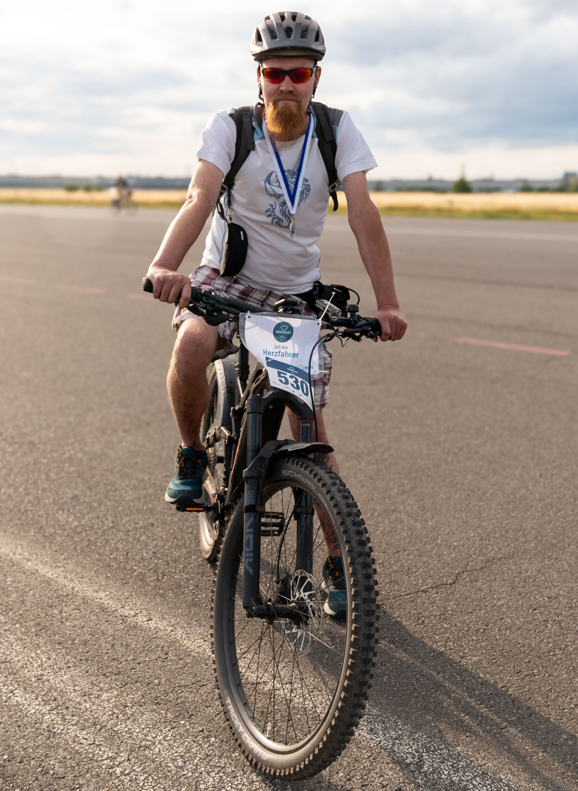 Ein Mann mit Bart, einer Sonnenbrille und einem T-Shirt sitzt auf einem Mountainbike. Er trägt eine Medaille um den Hals und hat eine Startnummer auf dem Fahrrad angebracht. Die Umgebung scheint eine asphaltierte Fläche zu sein, vermutlich eine Rennstrecke.