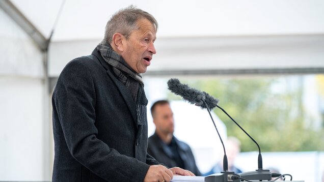 Ein Mann in einem schwarzen Mantel und mit einem Schal spricht leidenschaftlich am Rednerpult. Vor ihm steht ein Mikrofon, während im Hintergrund eine unscharfe Person zu sehen ist. Die Szene scheint bei einer Veranstaltung unter einem Zeltdach zu stattfinden.