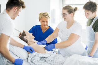 Fünf Personen stehen um ein Bett mit einer Übungspuppe, die medizinische Maßnahmen simuliert. Drei Personen führen unterschiedliche Handlungen am Oberkörper der Puppe aus, während eine Lehrkraft die Gruppe anleitet. Die Umgebung wirkt hell und modern, geeignet für medizinische Schulungen.