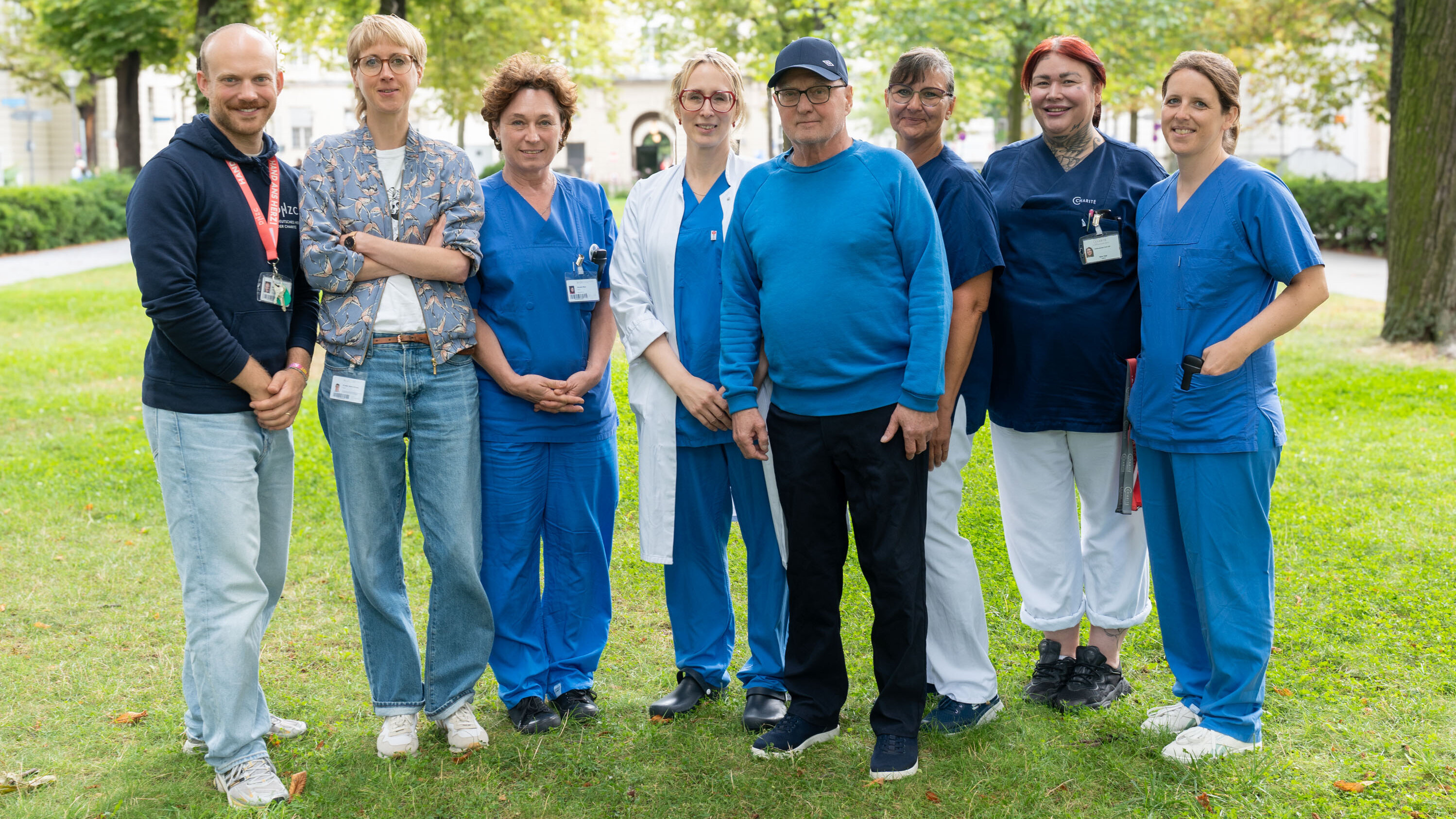 Eine Gruppe von acht Personen steht auf einer Wiese. Sie tragen verschiedene blaue Arbeitskleidung und wirken freundlich. Im Hintergrund sind Bäume und ein Park sichtbar. Die Personen scheinen in einem medizinischen oder pflegerischen Bereich tätig zu sein.