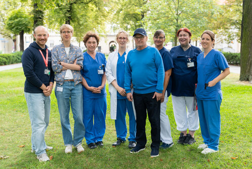 Eine Gruppe von acht Personen steht auf einer Wiese. Sie tragen verschiedene blaue Arbeitskleidung und wirken freundlich. Im Hintergrund sind Bäume und ein Park sichtbar. Die Personen scheinen in einem medizinischen oder pflegerischen Bereich tätig zu sein.