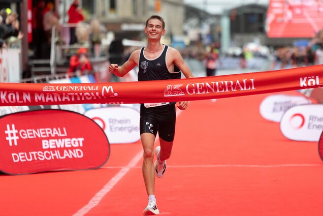 Ein strahlender Laufteilnehmer überquert die Ziellinie bei einem Marathon. Der Wettkampf ist durch ein rotes Band markiert, auf dem "Köln Marathon" steht. Der Athlet trägt ein dunkelblaues Trikot und lächelt, während er seine Arme in die Luft hebt.