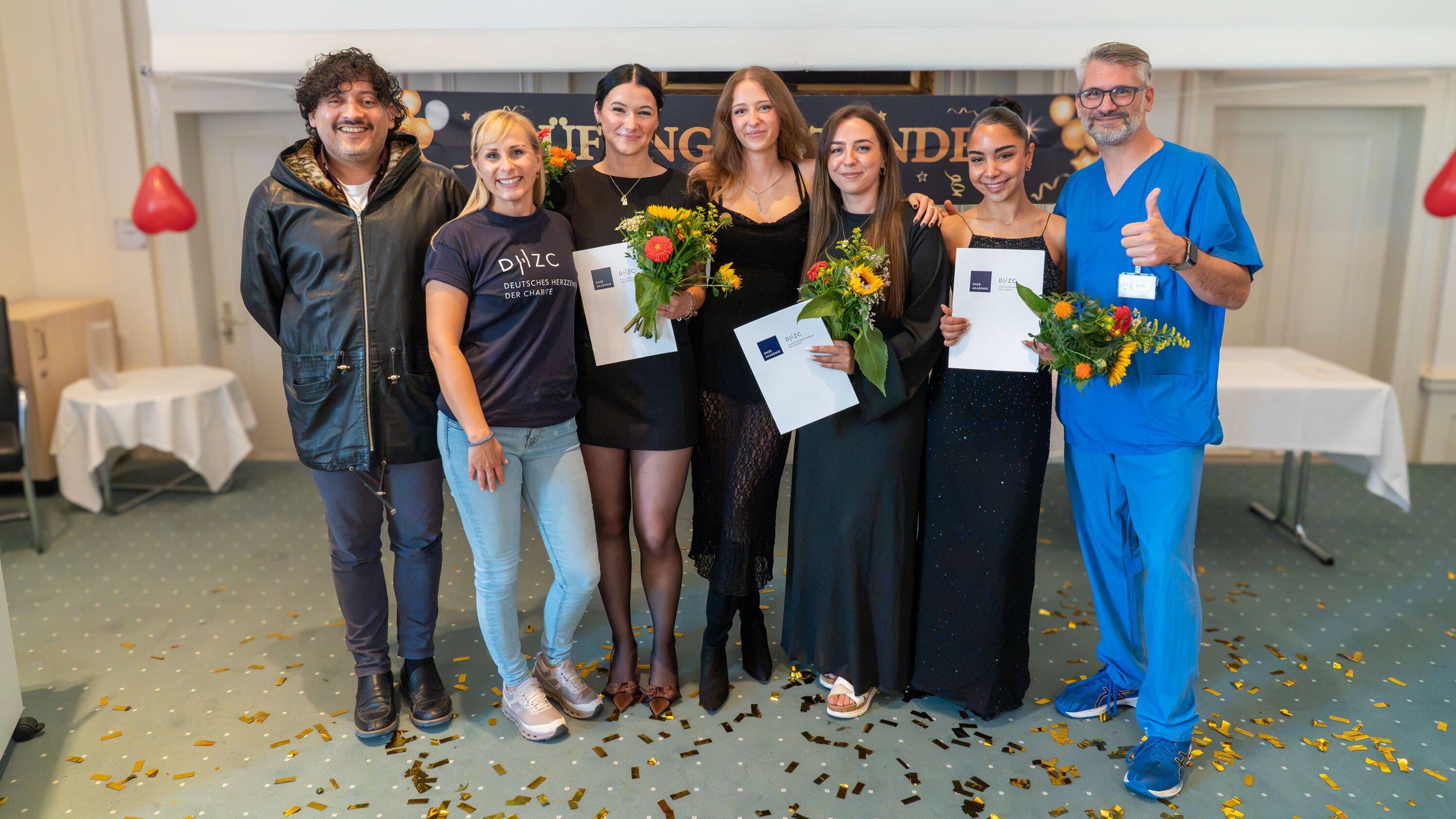 Sechs Personen posieren für ein Gruppenfoto in einem festlichen Raum. Alle halten Blumen und Zertifikate in den Händen. Die Stimmung ist feierlich und freundlich, passend zu einem Anlass wie einer Abschlussfeier oder einer Preisverleihung.