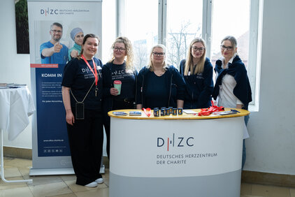 Sechs Frauen stehen an einem Empfangstisch des Deutschen Herzzeitungszentrums. Eine von ihnen hält einen Kaffeebecher. Im Hintergrund steht ein Banner mit Informationen über die Organisation. Sie lächeln und tragen einheitliche T-Shirts. Der Tisch ist mit Werbematerialien und einer roten Schleife dekoriert.