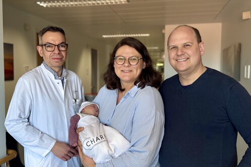 Ein Arzt in weißem Kittel steht neben einer lächelnden Frau, die ein Baby in einer Decke hält. Neben ihnen steht ein lächelnder Mann. Die Atmosphäre ist freundlich und feierlich, was auf die Geburt des Babys hindeutet.